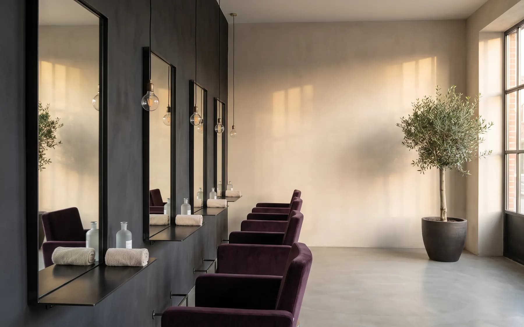 Symbolbild: Blick durch einen modernen Friseursalon — schmale schwarz gerahmte Spiegel an einer graphitfarbenen Wand, Stühle mit dunkelviolettem Samt, heller Putz und Tageslicht.
