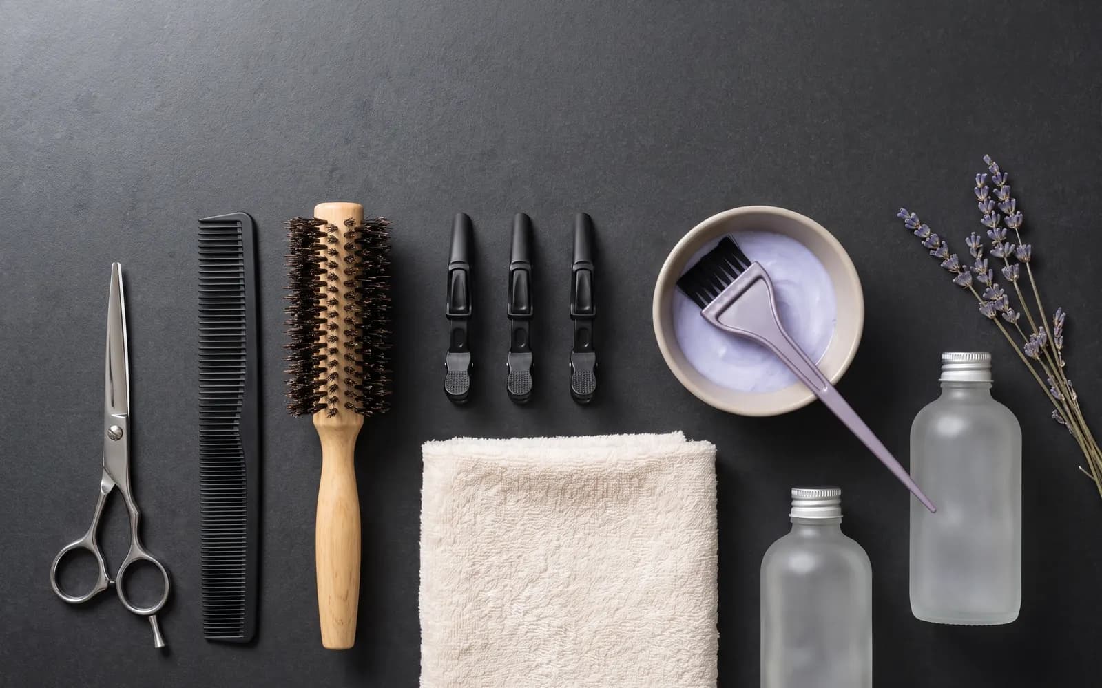 Symbolbild: Friseurwerkzeug von oben auf dunklem Stein — Schere, Kamm, Rundbürste, Klammern, ein Handtuch und eine Schale mit hellvioletter Tönungscreme.